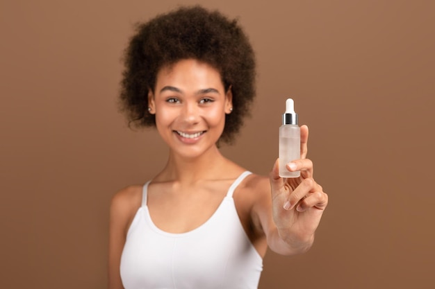 Smiling young african american curly female with perfect skin shows jar of serum recommends cosmetics