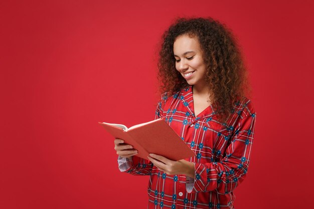 Smiling young african american girl in pajamas homewear posing while resting at home isolated on red background studio portrait. Relax good mood lifestyle concept. Mock up copy space. Reading book.