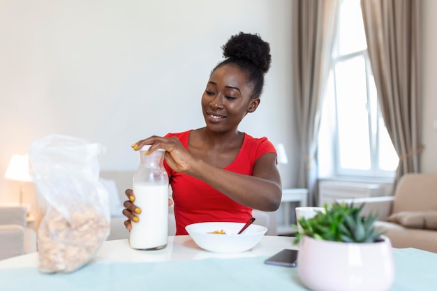 Smiling young African American woman pours corn flakes in plate with milk The girl has a healthy breakfast on stylish cozy home at the morning while checking her email on laptop