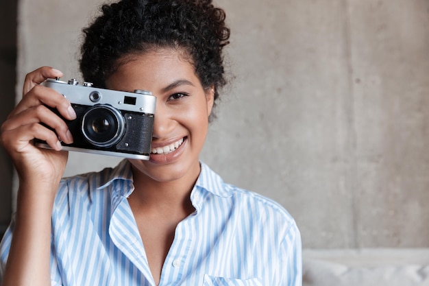 Smiling young african woman wearing shirt sitting on a couch at home, holding Portrait camera