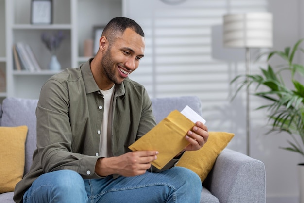 A smiling young africanamerican man is sitting on the sofa at home and opens an envelope in which he