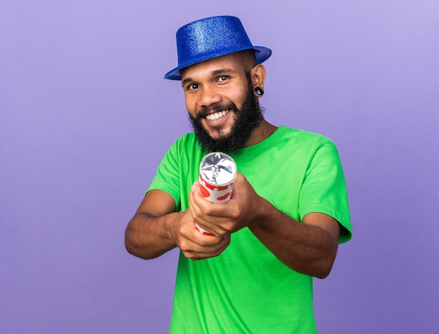 Smiling young afro-american guy wearing party hat holding out confetti cannon at front isolated on blue wall