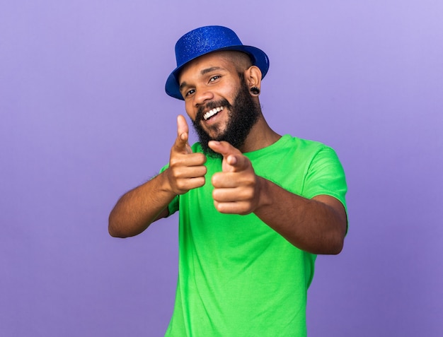 Smiling young afro-american guy wearing party hat points at camera 