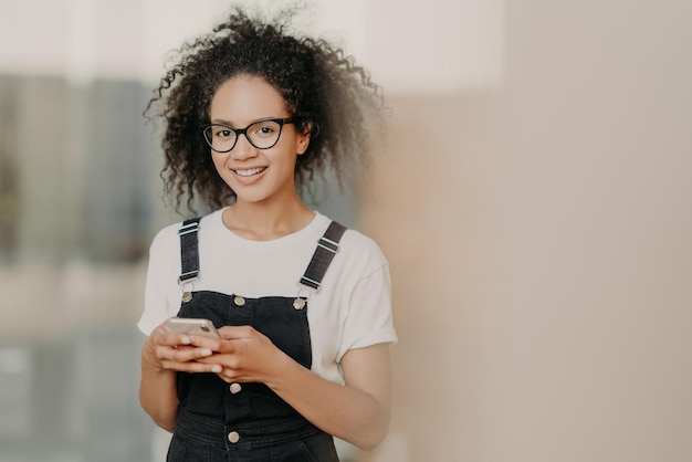 Smiling young Afro female holds modern cell phone uses mobile app lives in world of modern technologis addicted to social networks chats with friends online dressed in fashionable outfit