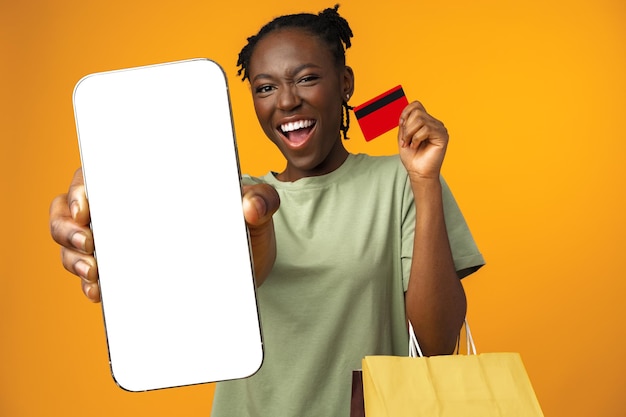 Smiling young afro girl holds smart phone and credit card in yellow studio