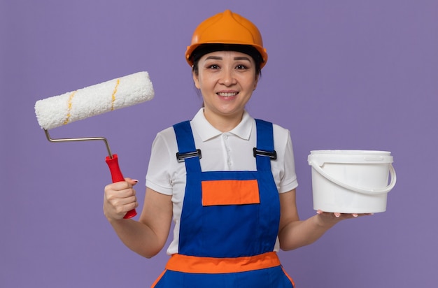 Smiling young asian builder woman with orange safety helmet holding paint roller and oil paint 