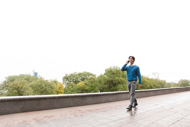 Smiling young asian business man talking on mobile phone while walking outdoors