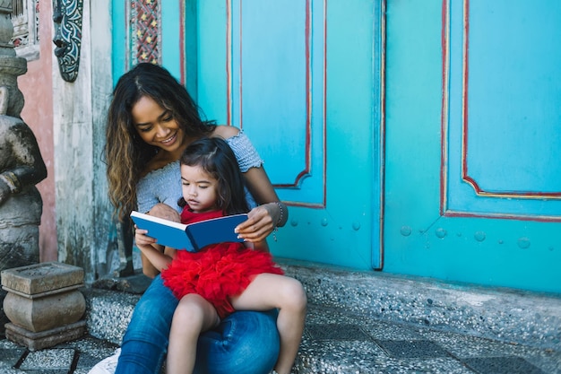 Smiling young Asian lady sitting with daughter on knees and reading book