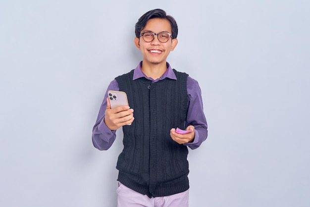 Smiling young Asian man in casual shirt using smartphone and holding money banknotes isolated on white background