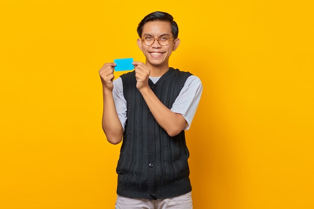 Smiling young Asian man holding credit card while looking at camera on yellow background