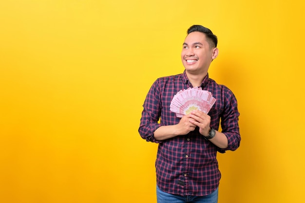 Smiling young Asian man holding money banknotes and looking at copy space isolated over yellow background Profit and wealth concept