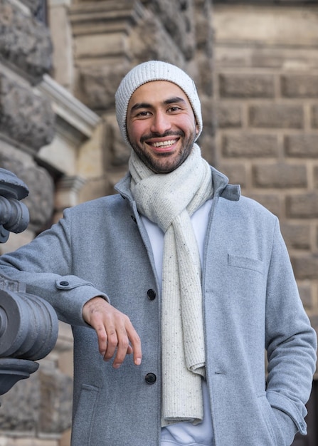 A smiling young Asian man in a light coat and a white knitted hat is traveling around the world Taking in the sights of a new place Closeup portrait