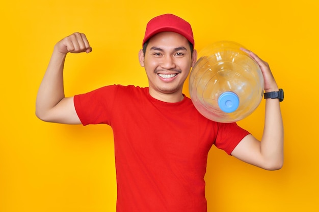 Smiling young Asian man in red cap tshirt uniform employee work as dealer courier holding water bottle showing muscle isolated on yellow background Professional Delivery service concept