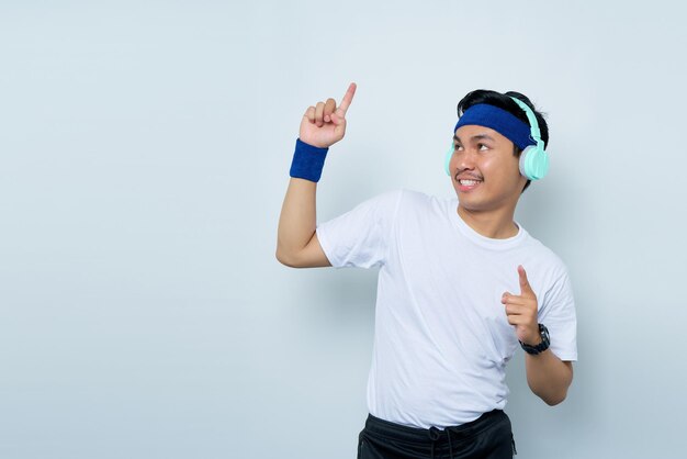 Smiling young Asian man sporty fitness trainer instructor in blue headband and white tshirt pointing finger at copy space isolated over white background