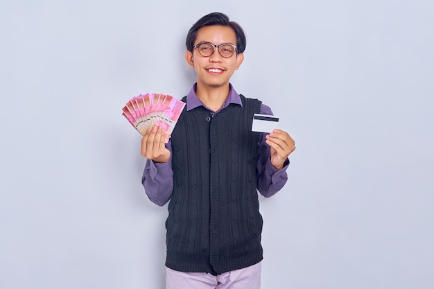 Smiling young Asian man in vest shirt showing money rupiah banknotes and credit card isolated on white background