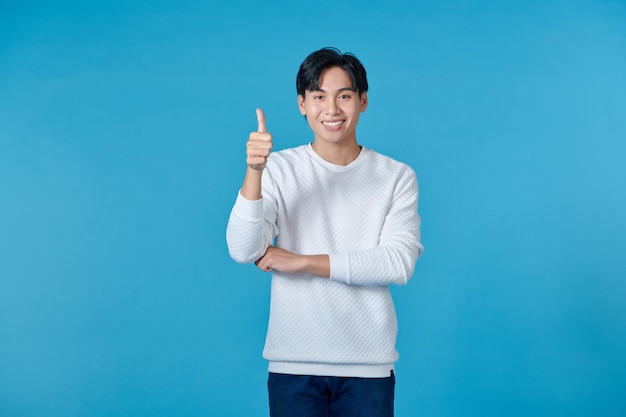 Smiling young asian man wearing white jumper and jeans on blue background giving thumbs up