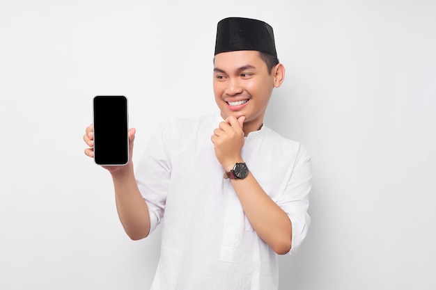 Smiling young Asian Muslim man showing smartphone screen touching his chin thinking about the offer on white background People religious Islam lifestyle concept celebration Ramadan and ied Mubarak