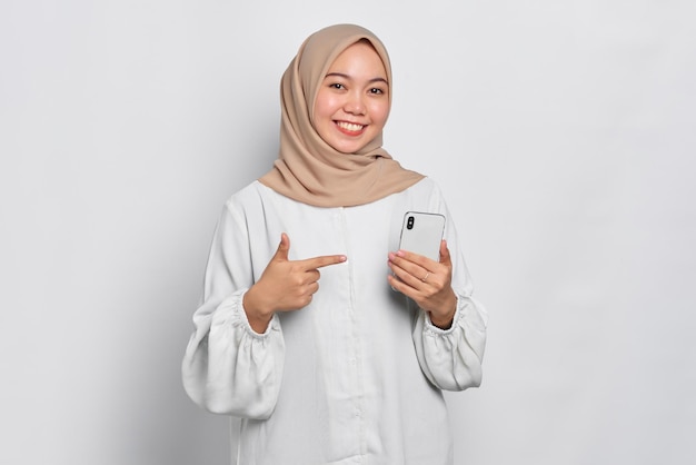 Smiling young Asian Muslim woman pointing fingers at mobile phone isolated over white background