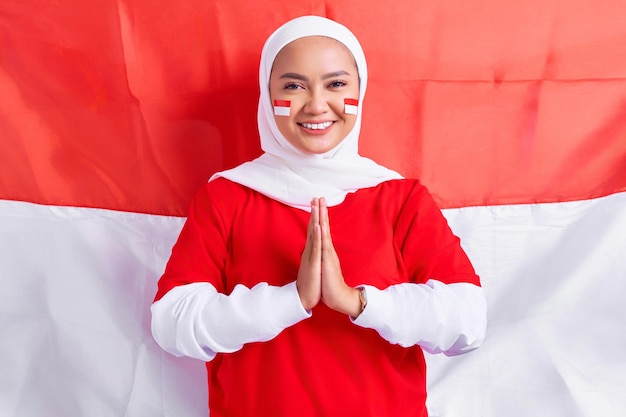 Smiling young Asian muslim woman in red white tshirt greeting to celebrating Indonesia independence day on August 17 isolated on flag indonesian background