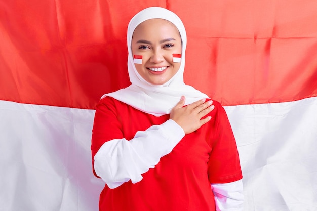 Smiling young Asian muslim woman in red white tshirt greeting to celebrating Indonesia independence day on August 17 isolated on flag indonesian background