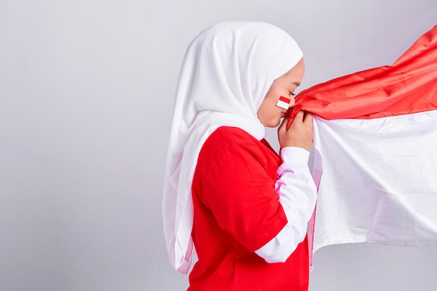 Smiling young Asian muslim woman in red white tshirt respect or salute gesture while kiss flag indonesian and celebrating indonesian independence day August 17 isolated on white background