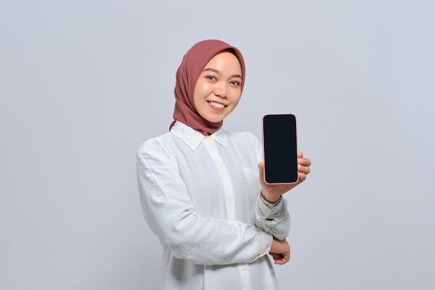 Smiling young Asian Muslim woman showing mobile phone blank screen recommending new App isolated over white background