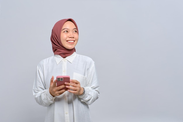 Smiling young Asian Muslim woman using mobile phone and looking away at copy space isolated over white background