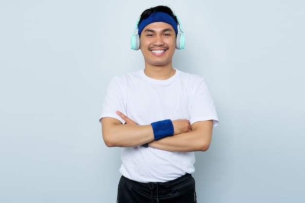 Smiling young Asian sportman in blue headband and sportswear white tshirt while listening music favorite with headphones crossed arms chest and looking confident isolated on white background