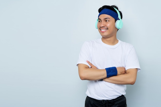 Smiling young Asian sportman in blue headband and sportswear white tshirt while listening music favorite with headphones crossed arms chest and looking confident isolated on white background