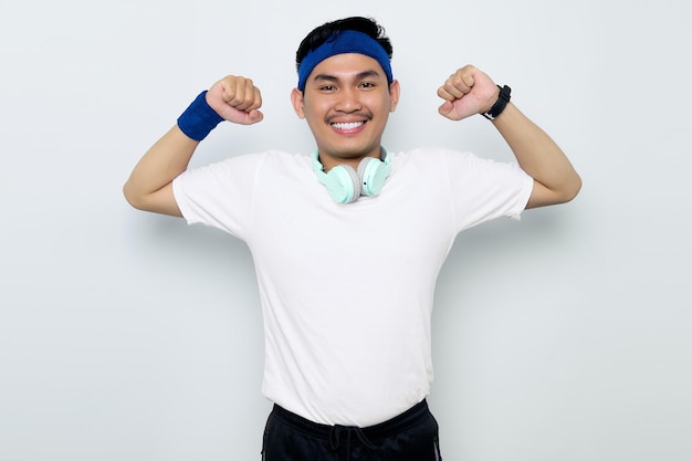 Photo smiling young asian sportman in blue headband and white tshirt with headphones raises arms and shows biceps demonstrated her achievement after training in a gym isolated on white background