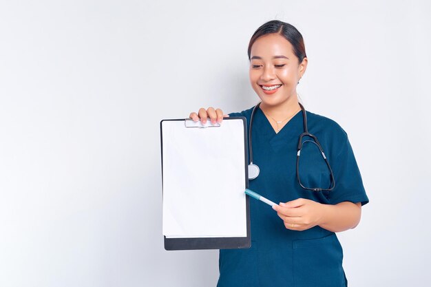 Photo smiling young asian woman nurse wearing blue uniform with a stethoscope hold clipboard and pen explains info on the clipboard pointing paper isolated on white background healthcare medicine concept