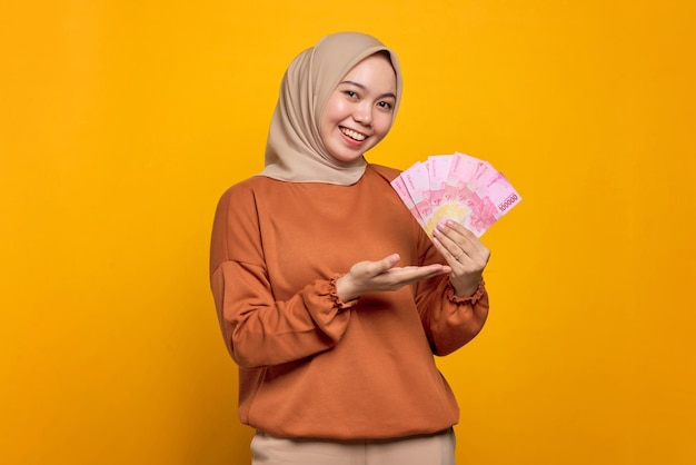 Smiling young Asian woman in orange shirt showing money banknotes isolated over yellow background