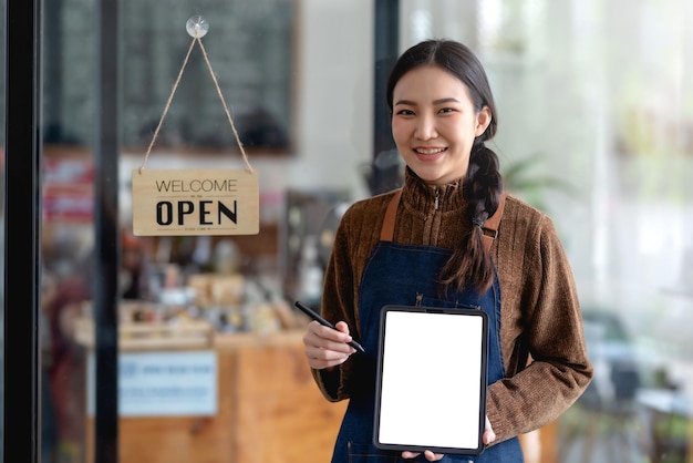 Smiling young Asian woman owner standing holding a tablet blank white screen and open sign board Looking at camera