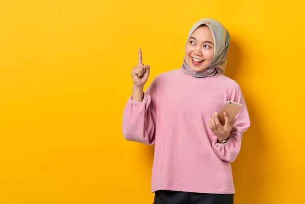 Smiling young Asian woman in pink shirt holding mobile phone and pointing fingers up having a good idea on yellow background