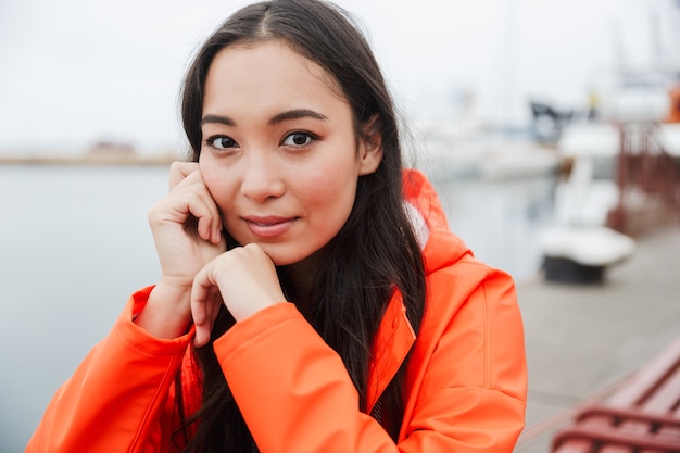 Smiling young asian woman wearing raincoat spending time outdoors walking at the coastland, sitting on a bench