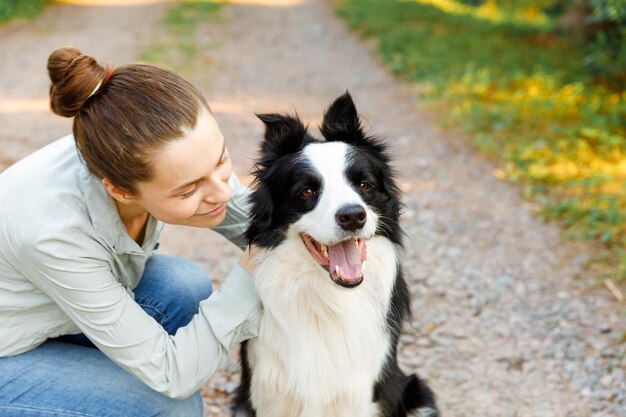 Smiling young attractive woman playing with cute puppy dog border collie on summer outdoor background. Girl holding embracing hugging dog friend. Pet care and animals concept.