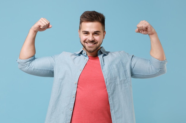 Smiling young bearded guy 20s in casual shirt posing isolated on pastel blue wall background studio portrait. People sincere emotions, lifestyle concept. Mock up copy space. Showing biceps, muscles.