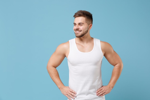 Smiling young bearded guy 20s in white singlet posing isolated on pastel blue background. Sport fitness healthy lifestyle concept. Mock up copy space. Standing with arms akimbo on waist looking aside.