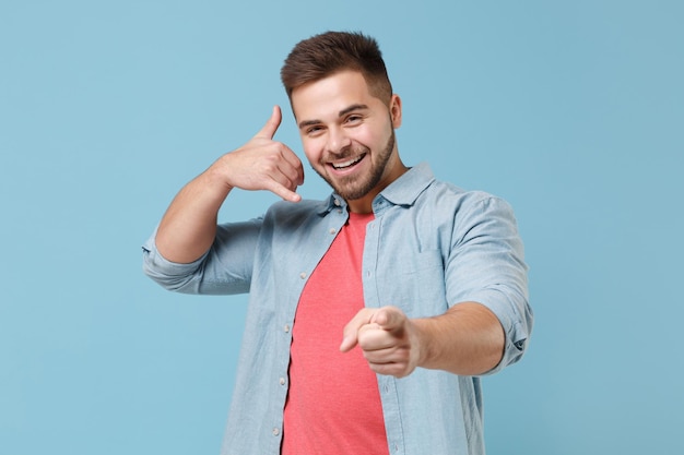 Photo smiling young bearded guy in casual shirt posing isolated on pastel blue background. people lifestyle concept. mock up copy space. doing phone gesture says call me back, point index finger on camera.