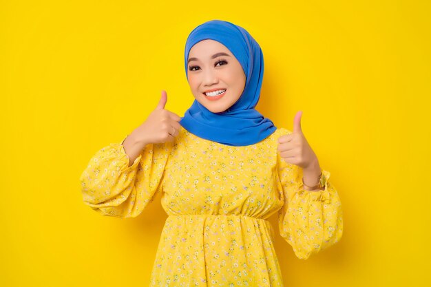 Smiling young beautiful Asian Muslim woman showing thumbs up isolated over yellow background