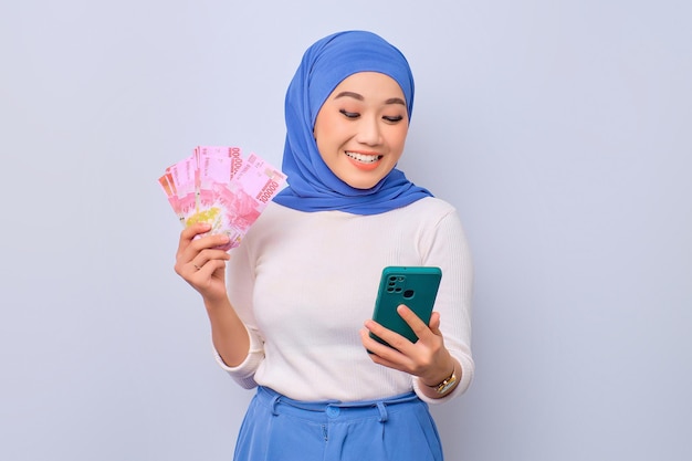 Smiling young beautiful Asian Muslim woman using mobile phone and holding money banknotes isolated over white background