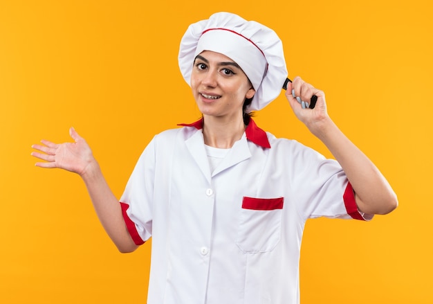 Smiling young beautiful girl in chef uniform holding frying pan behind head isolated on orange wall