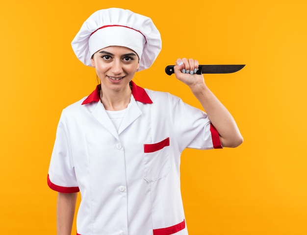 Smiling young beautiful girl in chef uniform holding knife isolated on orange wall