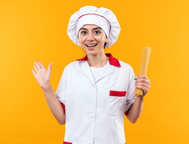 Smiling young beautiful girl in chef uniform holding spaghetti points with hand at side isolated on orange wall with copy space