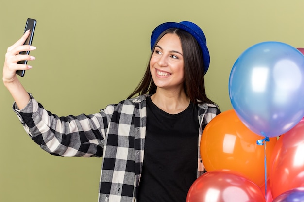 Smiling young beautiful girl wearing blue hat standing nearby balloons and take a selfie 