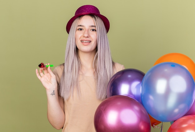 Smiling young beautiful girl wearing dental braces with party hat standing nearby balloons holding party whistle isolated on olive green wall