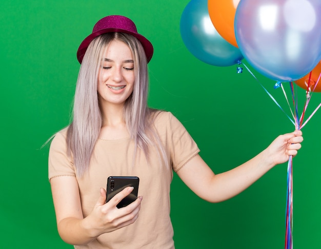 Smiling young beautiful girl wearing party hat and braces holding balloons looking at phone in her hand isolated on green wall