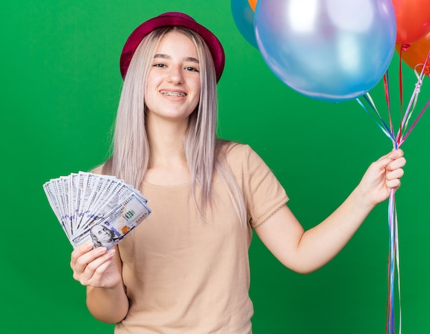 Smiling young beautiful girl wearing party hat holding balloons holding cash 