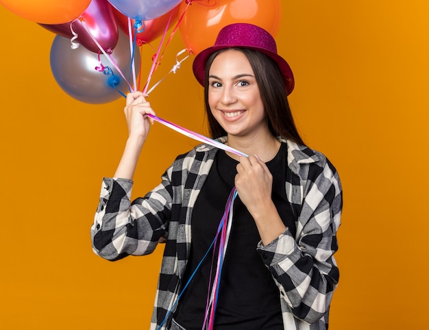 Smiling young beautiful girl wearing party hat holding balloons isolated on orange wall