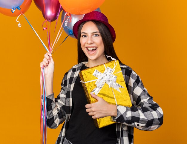 Smiling young beautiful girl wearing party hat holding balloons with gift box isolated on orange wall